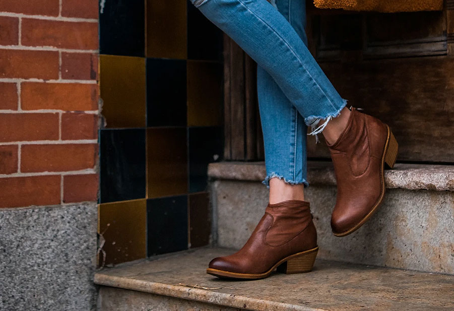 Woman wearing a pair of Aisley heeled booties in brown standing ion a concrete step.